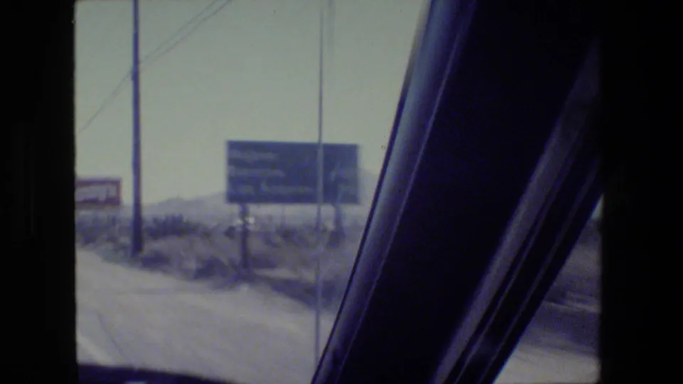 A moving 8mm view through the windshield shows blue roadside signs, a windshield pillar, and arid roadside terrain in California.