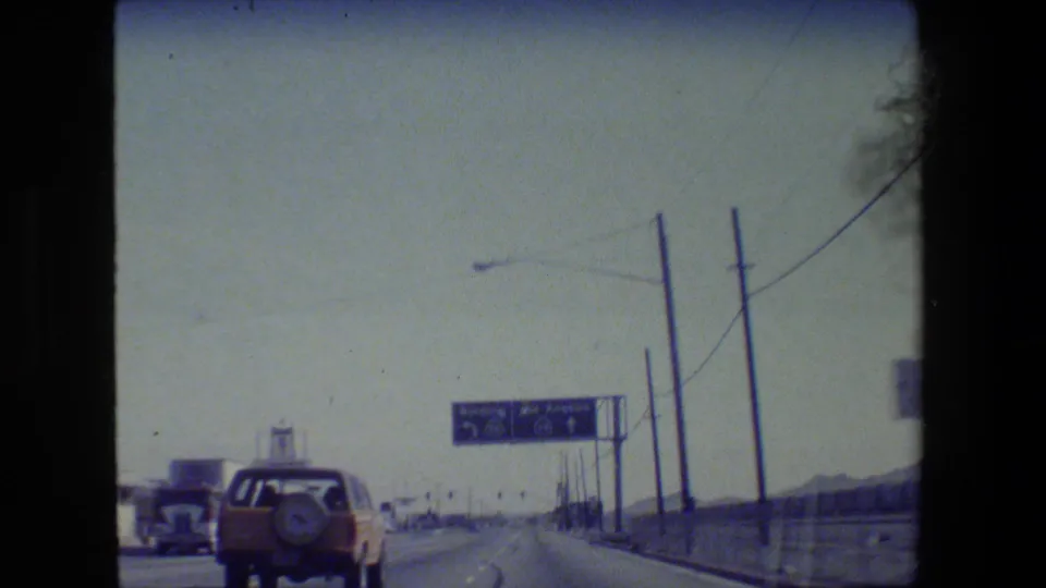 1984 California road-trip footage shows a car ahead on a highway approaching blue directional signage in dry terrain.