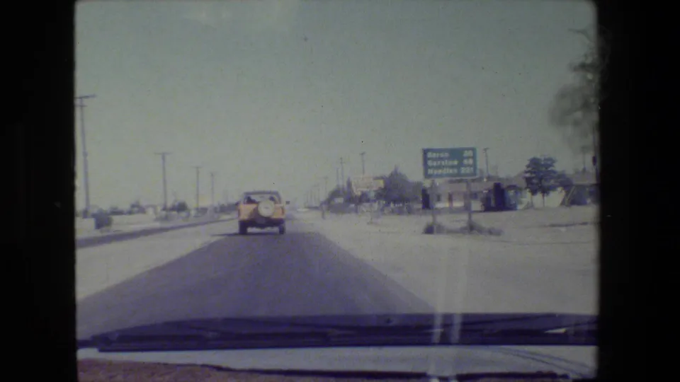 From inside the vehicle, the 8mm frame shows a car ahead and a blue highway sign listing Barstow and Los Angeles.