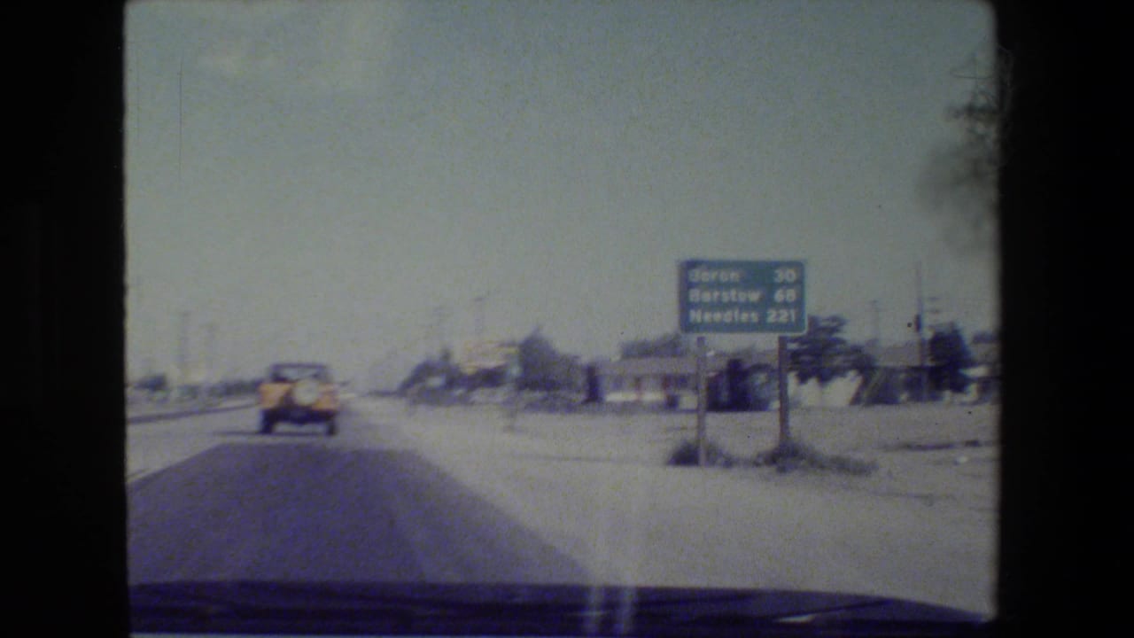 1984 8mm road-trip view through a windshield, showing a highway, dashboard edge, car ahead, and distant California road sign.