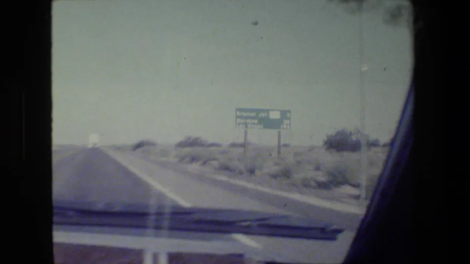 Home-movie windshield view of a straight California highway with arid roadside terrain and a guide sign ahead on the right.