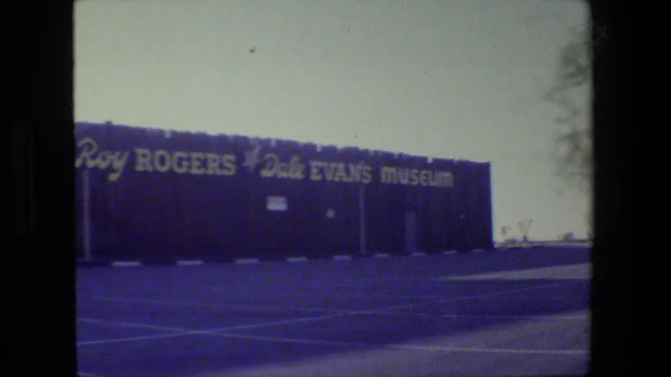 1984 8mm view of the Roy Rogers-Dale Evans Museum exterior, with large lettering across a dark building and parking lines in front.
