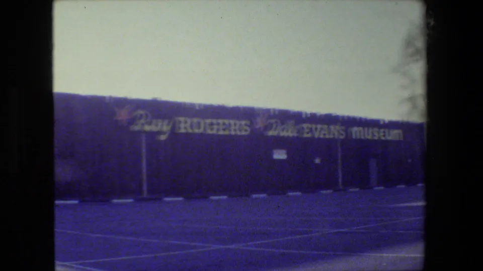 Visitor-shot 8mm frame of the Roy Rogers-Dale Evans Museum facade, with large name signage and empty parking spaces visible.