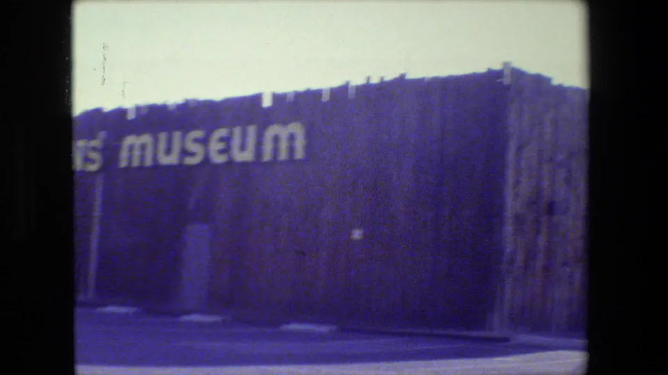 Super 8 frame with a partial view of the museum facade and large white lettering across a dark wall, filmed from outside.