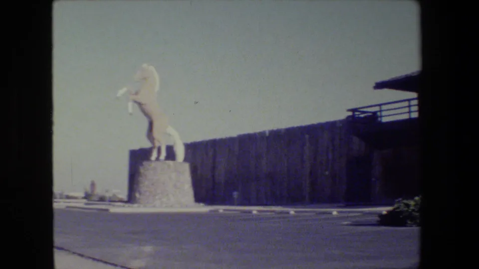 Super 8 frame showing the white horse monument on its pedestal outside the museum, with pavement and dark exterior wall behind it.