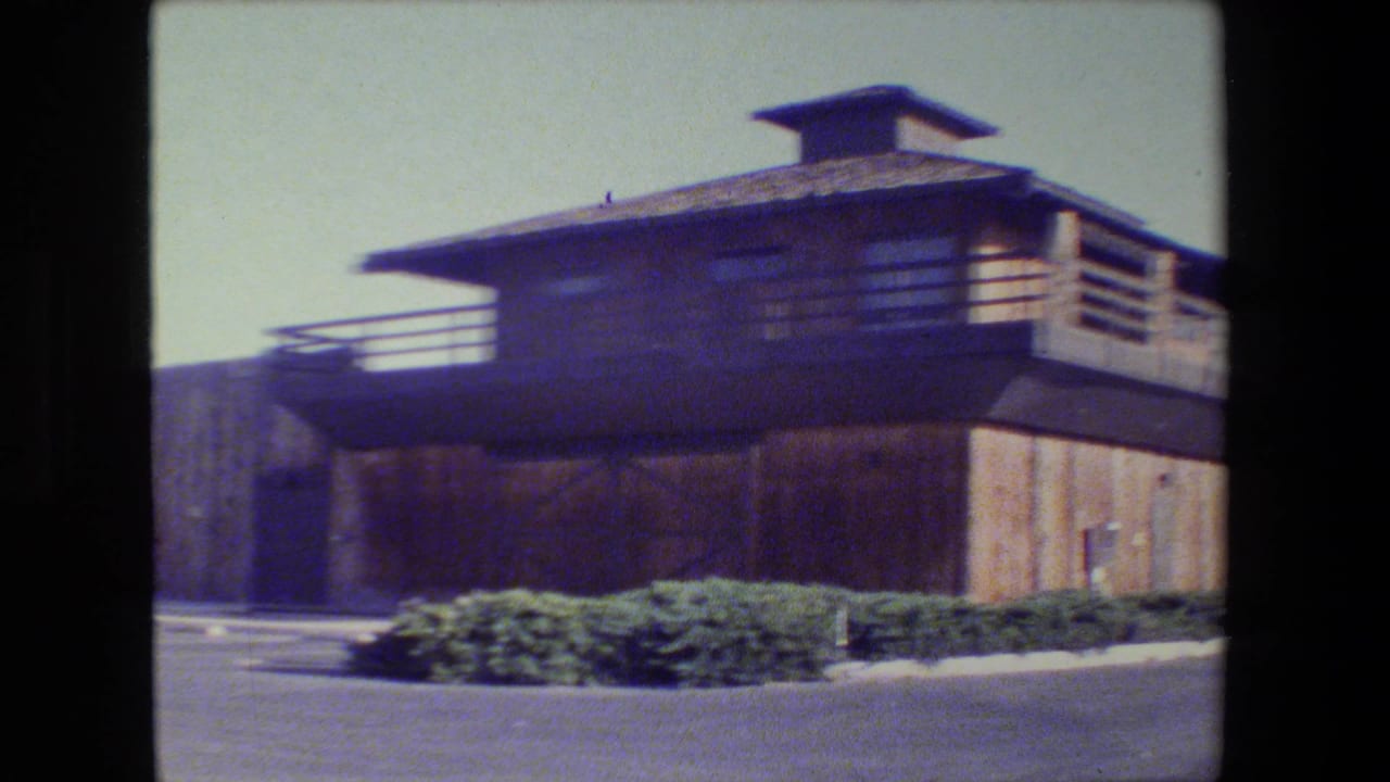 Two-story wood building with balcony railing, shrubs, asphalt, and bright sky.