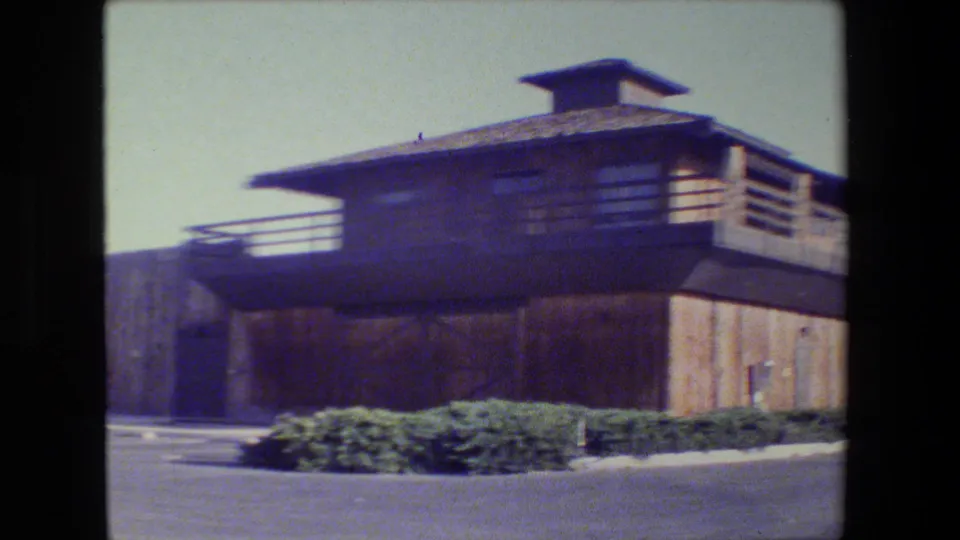 1984 San Francisco frame showing a two-story wooden building with upper deck railings, low shrubs, pavement, and strong daylight.