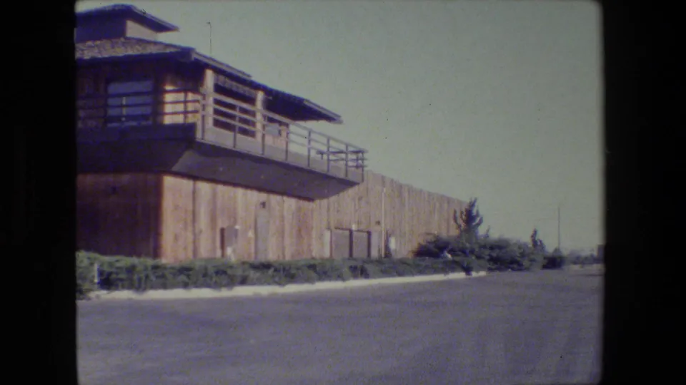 1984 San Francisco frame showing the building receding along a long wall, with deck railing, shrubs, pavement, and open sky at the end of the pan.