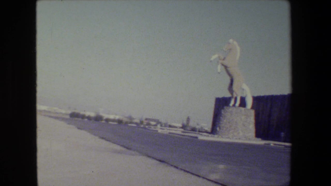 White rearing horse statue on a stone pedestal beside a road and dark fence.
