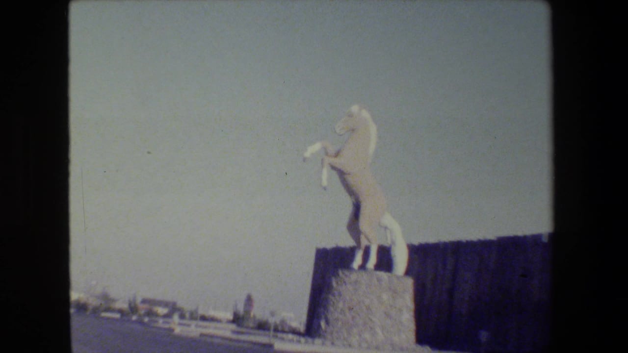 Rearing horse statue on a pedestal with roadway, fence, and pale sky around it.