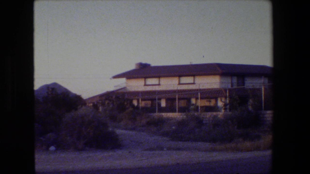 Two-story house with a dark roof, pale walls, shrubs, and a low wall beside a road.