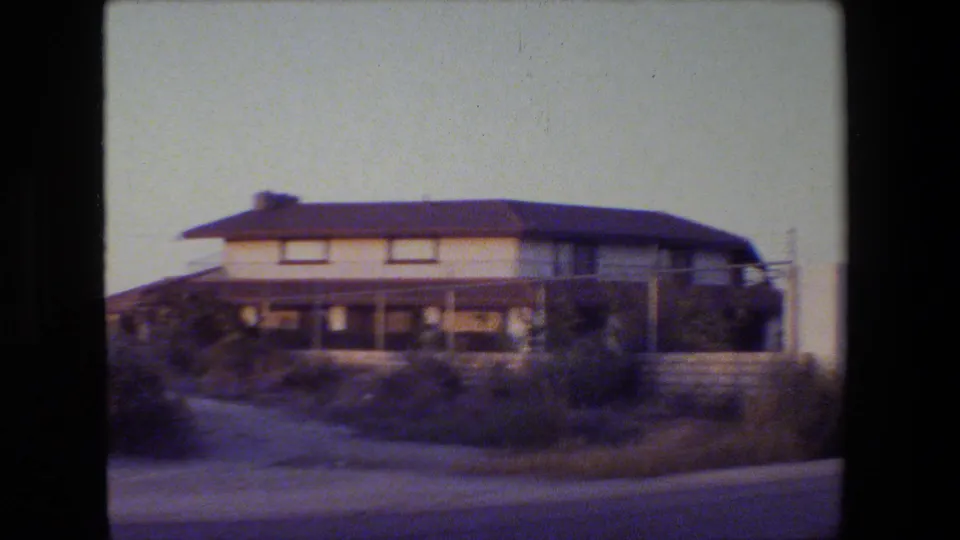 Closer 8mm drive-by frame of a residential house exterior, roadside curb, bushes, and low wall in the supplied San Francisco clip.