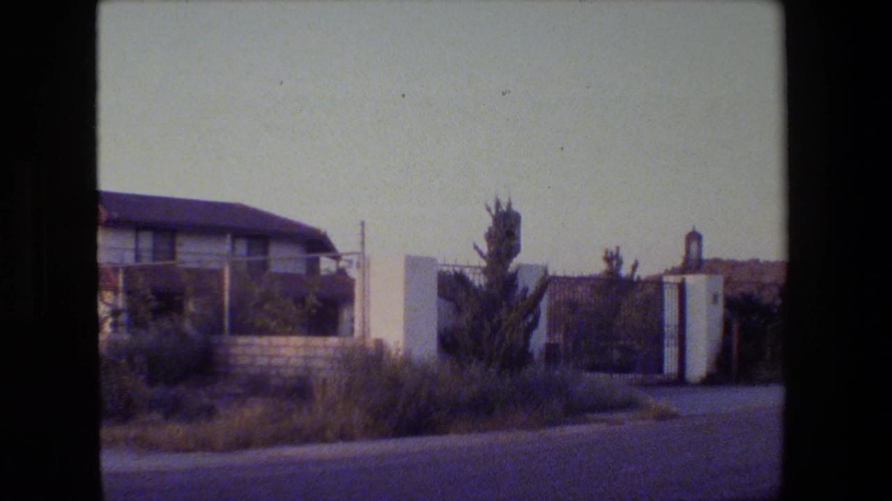 1984 home-movie frame of a gated California residential entrance with white pillars, shrubs, dry grass, and a two-story house.