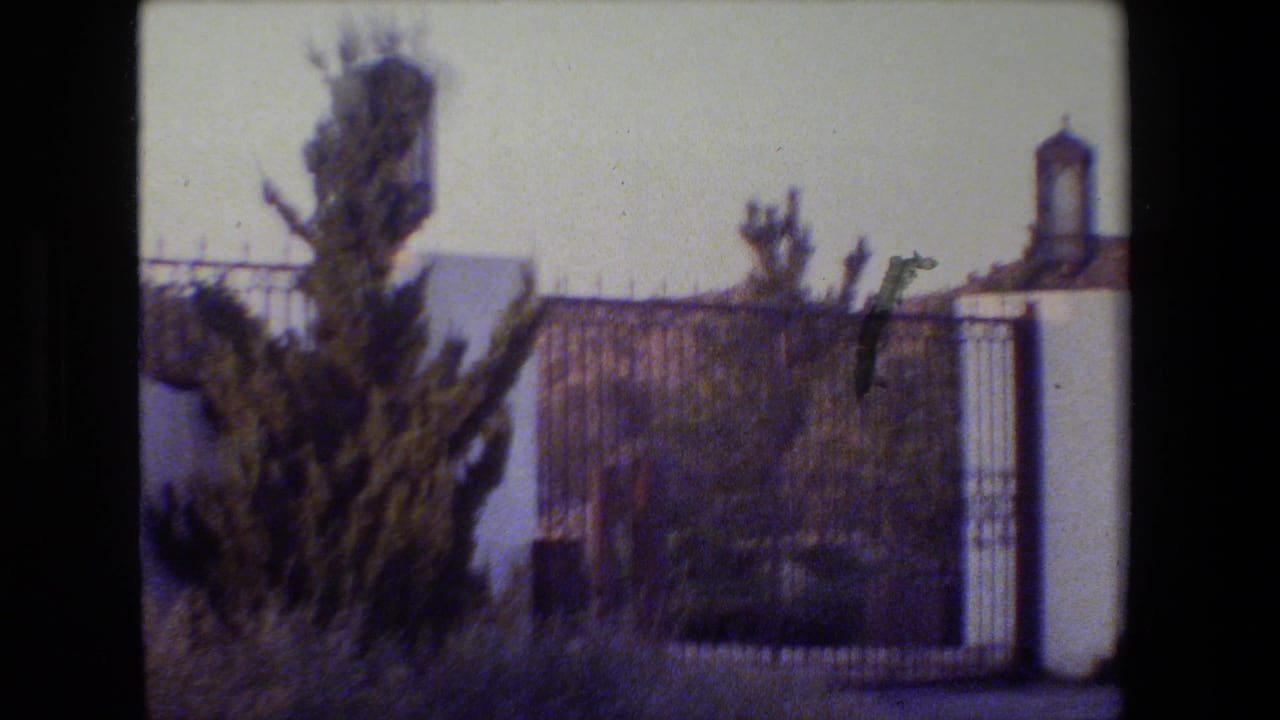 Close view of white gate pillars, iron fencing, shrubs, and hills beyond.