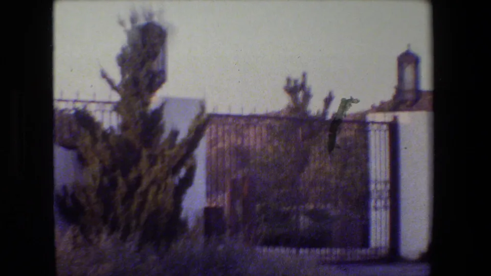 Closer 8mm view of the estate entrance with white pillars, wrought iron fencing, shrubs, and distant rolling hills.