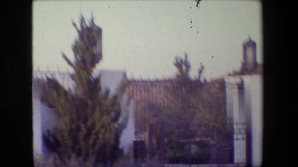 Soft 8mm frame showing the gated residential entrance, shrubs, white wall, and lantern-topped pillar in California.