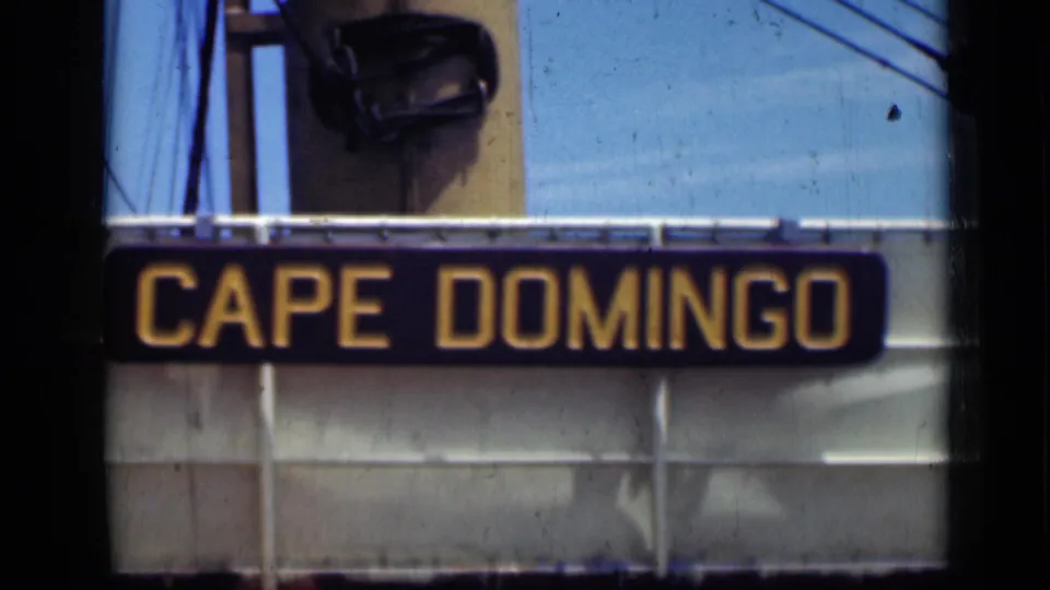 1946 8mm frame showing a black Cape Domingo sign with yellow lettering mounted on a pale structure, with film gate edges visible.