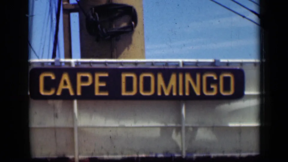 Close 8mm view of the Cape Domingo sign, its yellow letters centered against a dark board on a pale facade or ship structure.