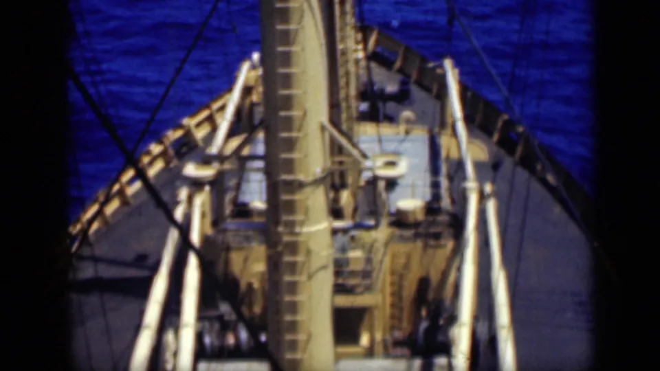 1946 8mm frame looking down from a ship mast toward the deck ladder, rigging lines, railings, and surrounding ocean water.