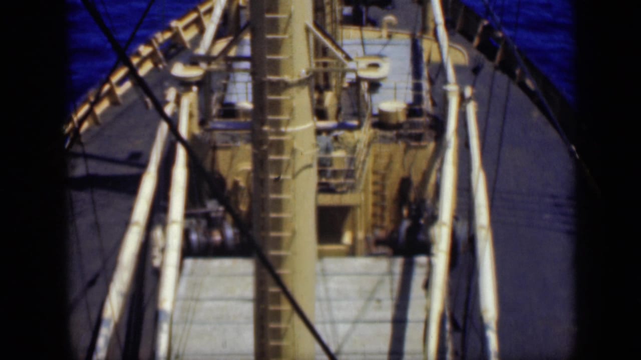 High-angle view of a ship deck with a tall mast, rigging, and blue water beyond.