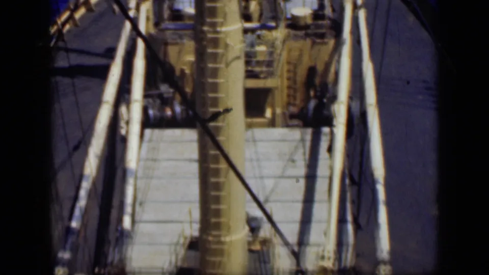 1946 8mm frame of a vessel underway, showing the deck, central mast, rigging, and surrounding blue water through dark borders.