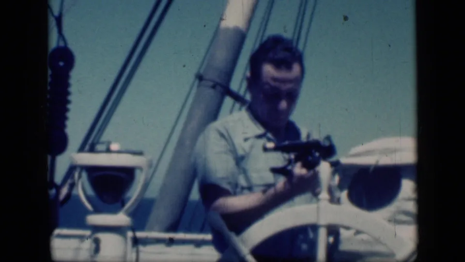 In this 1946 Cairo home-movie frame, a man holds a cine camera at chest height beside the sailboat wheel, mast, and rigging.