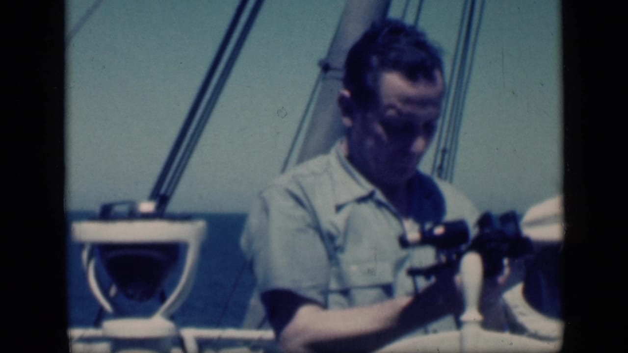 Man seated or standing on deck adjusts binoculars beside boat hardware.