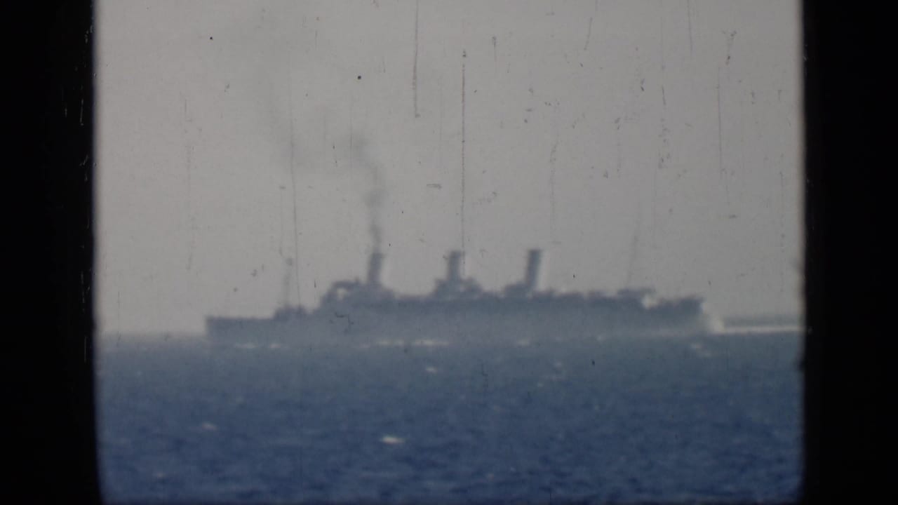 Large ship seen far offshore with smoke and a white wake.