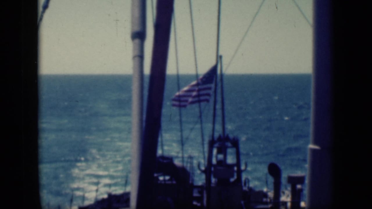1946 home-movie frame showing an American flag on a ship deck at sea, with rigging, mast lines, wake, and blue horizon visible.