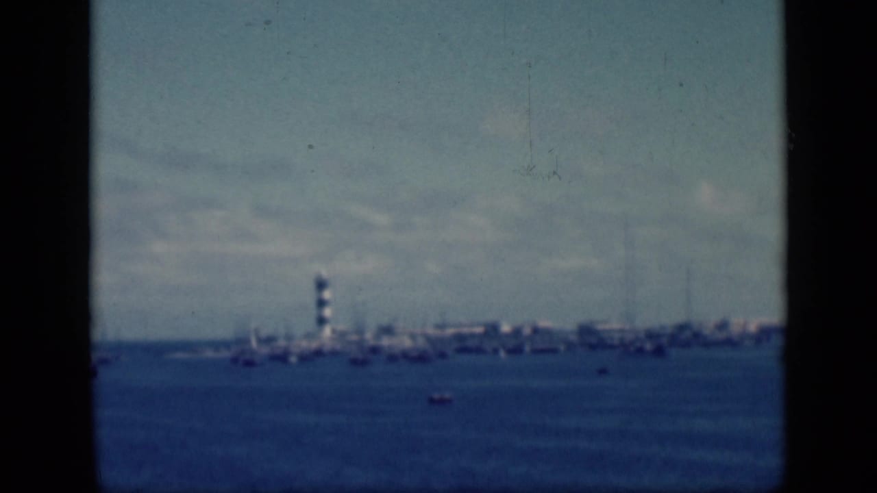 1946 Egypt 8mm frame showing a distant striped lighthouse on a shoreline with small boats on blue harbor water.