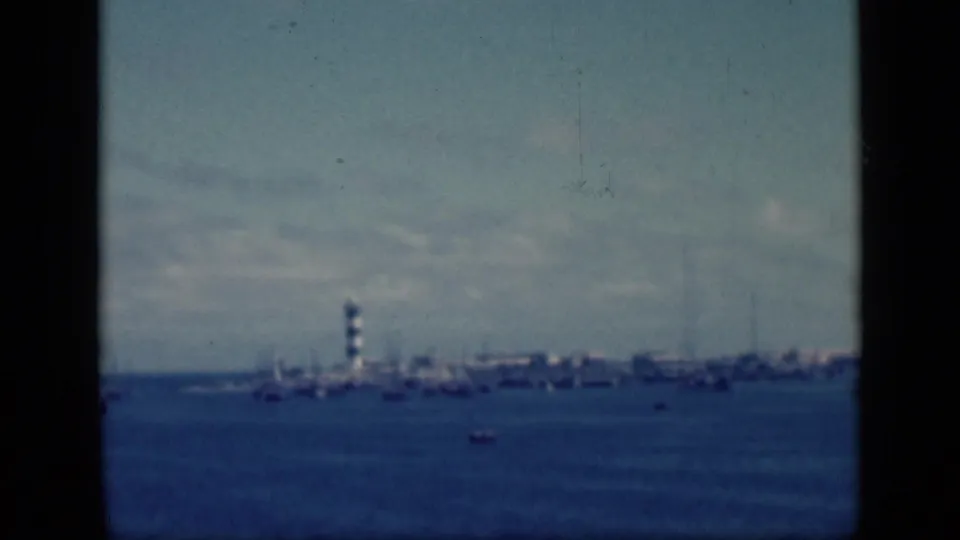 1946 Egypt 8mm frame showing a distant striped lighthouse on a shoreline with small boats on blue harbor water.