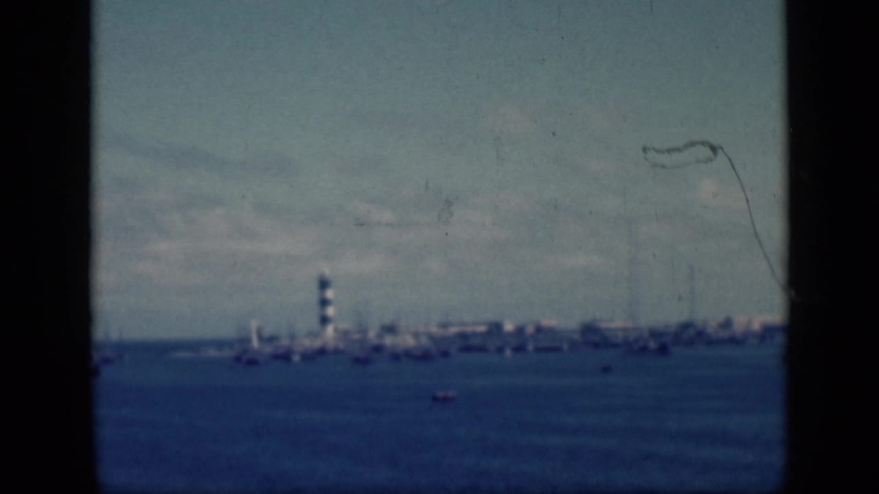 1946 Egypt home-movie frame with a distant lighthouse and harbor shoreline viewed across blue water through dark film borders.