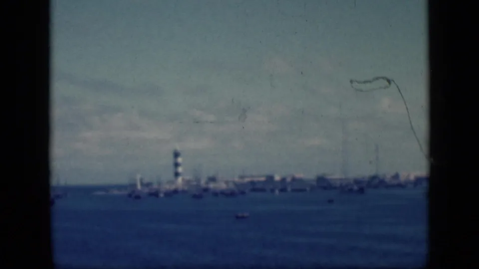 1946 Egypt home-movie frame with a distant lighthouse and harbor shoreline viewed across blue water through dark film borders.