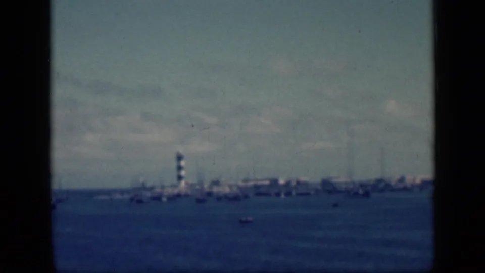 Blue-tinted 1946 Egypt 8mm frame showing a far shoreline lighthouse, scattered boats, and low waterfront structures.
