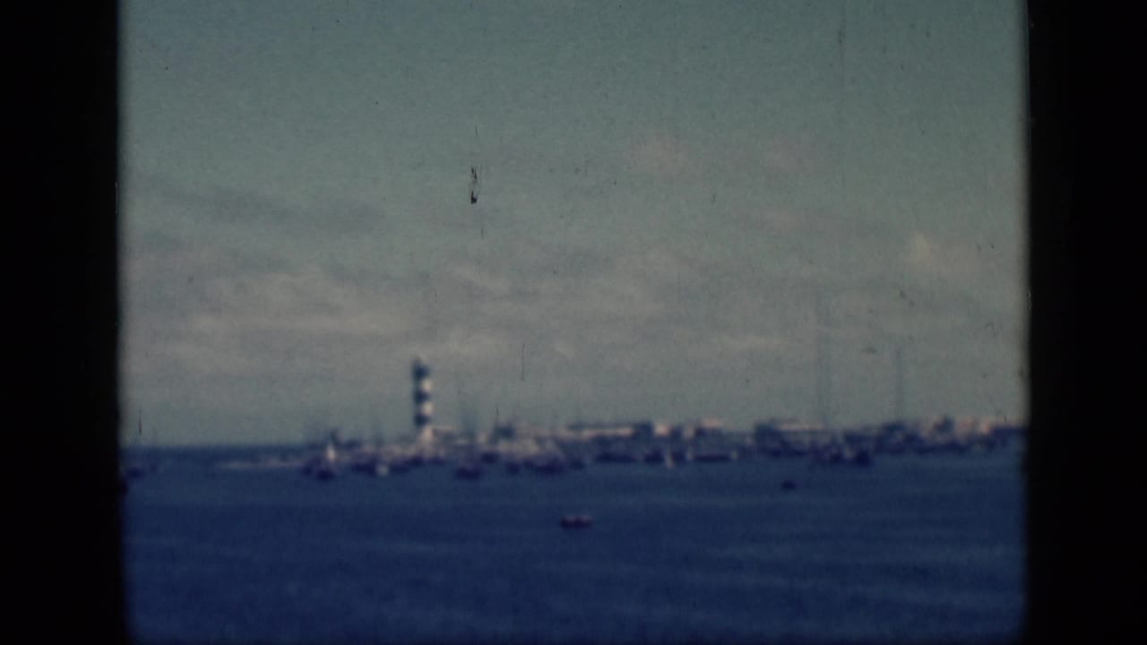 1946 Egypt home-movie frame showing a distant harbor across water, with a small striped lighthouse and waterfront shapes under a hazy sky.