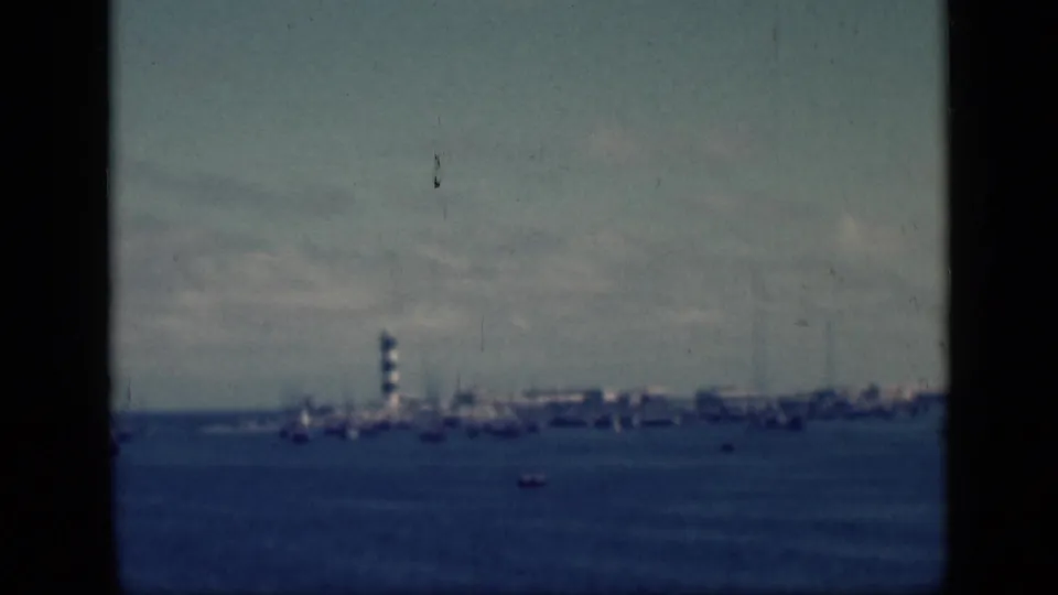 1946 Egypt home-movie frame showing a distant harbor across water, with a small striped lighthouse and waterfront shapes under a hazy sky.