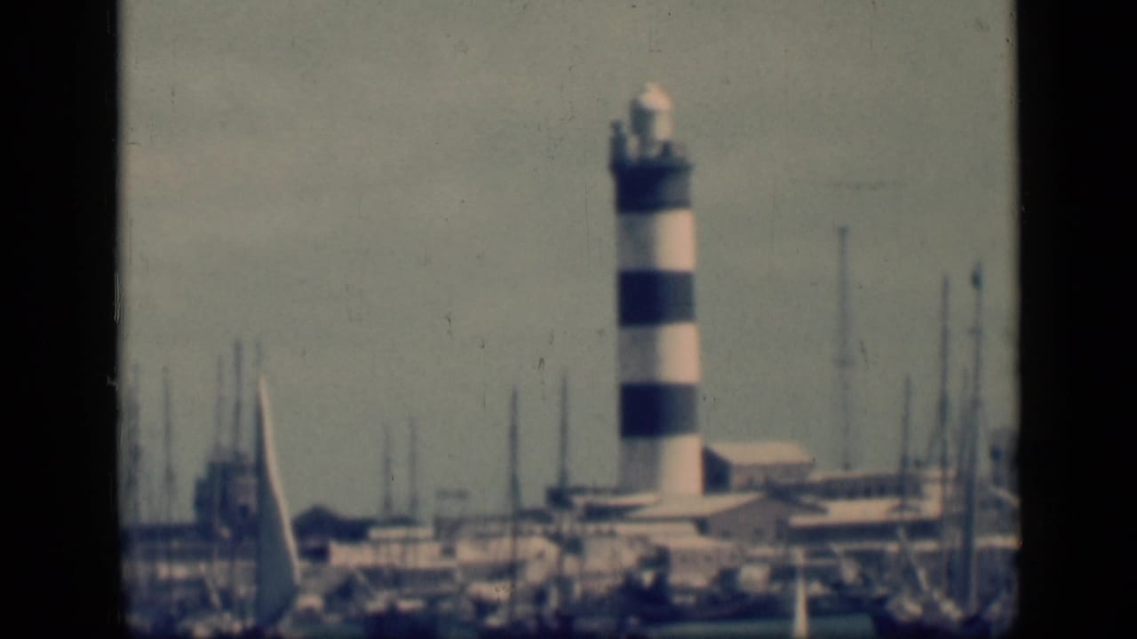 Closer view of a black-and-white striped lighthouse beside marina masts and low buildings.