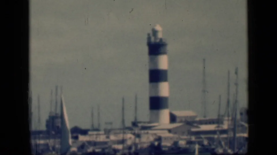 1946 Egypt home-movie still showing the striped lighthouse in soft focus, framed by sailboat masts and waterfront structures.