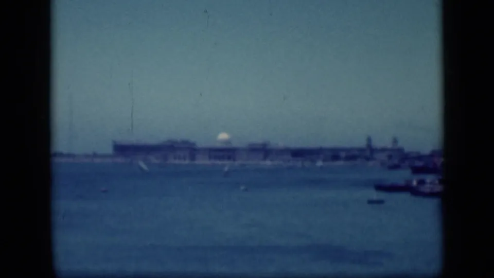 1940s Egypt frame with a long waterfront building on the horizon, small boats on the water, and a dark tower-like structure nearby.