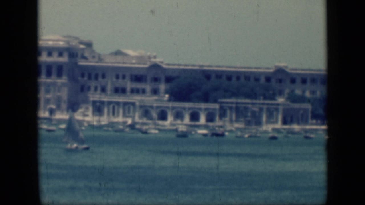 1946 Cairo waterfront view with a small sailboat crossing blue harbor water before a long pale building and arched colonnades.