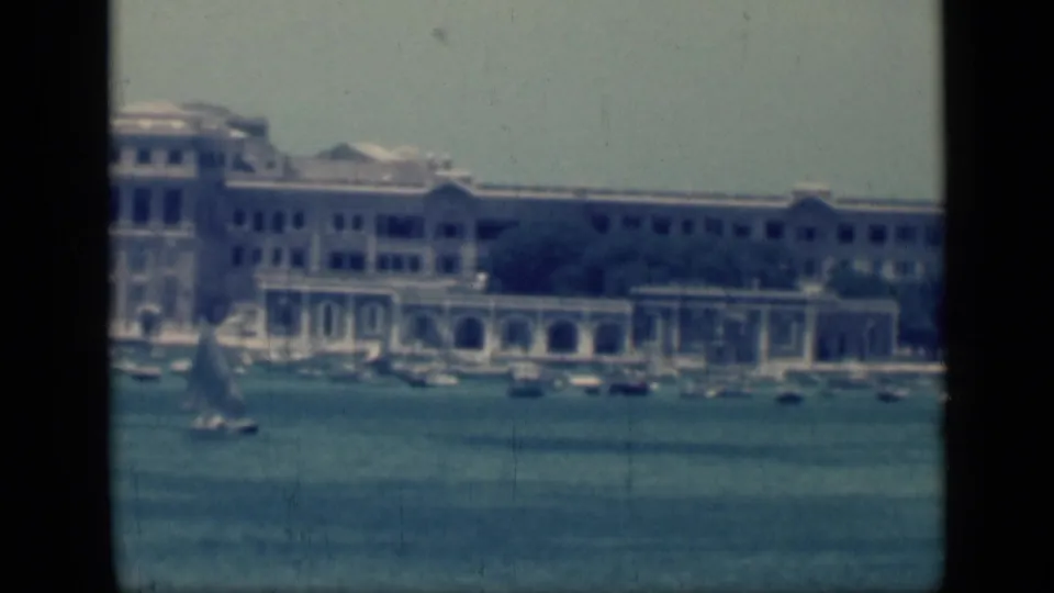 1946 Cairo waterfront view with a small sailboat crossing blue harbor water before a long pale building and arched colonnades.