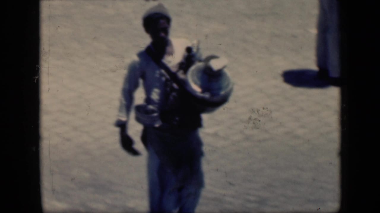 Man in light clothing walking across a sunlit paved courtyard carrying a metal container.