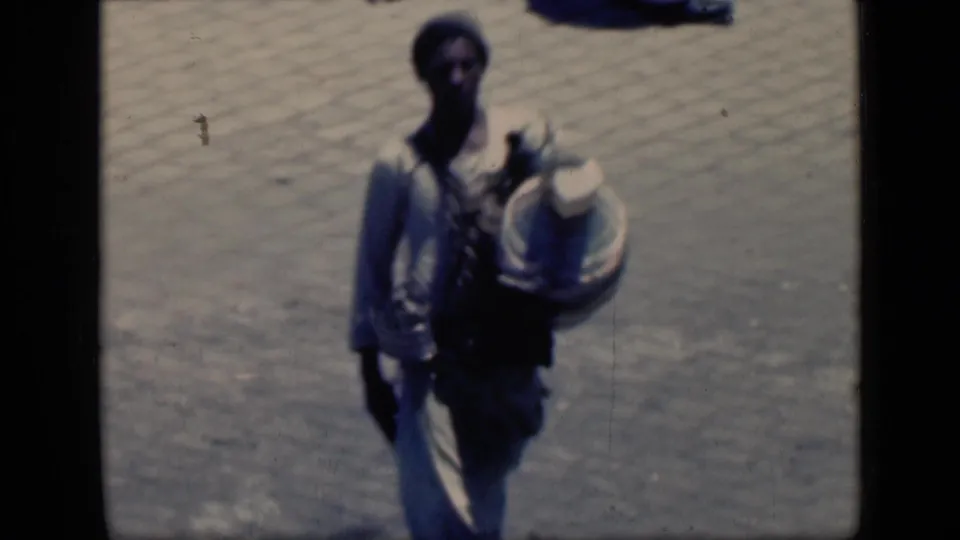 1946 Cairo home-movie frame with an adult man near the camera, carrying a large round metal container over brick paving.