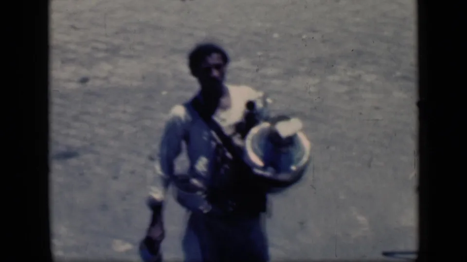 1946 Cairo frame showing an adult man in light clothing with a shoulder strap or load, carrying a large metal container in harsh sun.