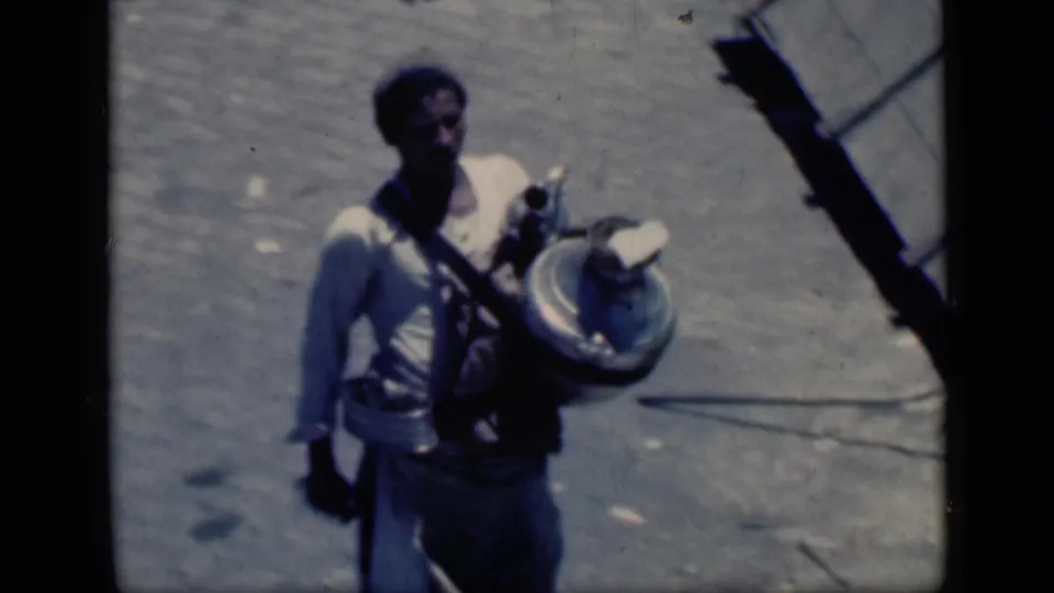 In 1946 Cairo home-movie footage, a young man in a light shirt walks over dusty ground carrying a large round object.