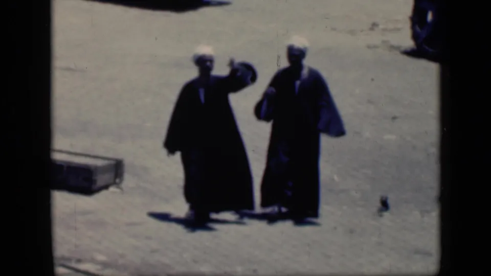 Two men in dark robes and white head coverings walk side by side across a sunlit Cairo open area in 1946 footage.