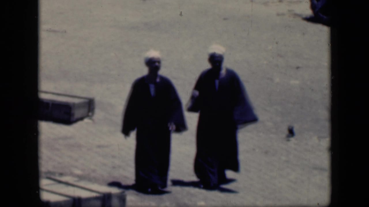 In 1946 Cairo footage, two adults in dark robes and white head coverings walk together across sunlit dusty ground.