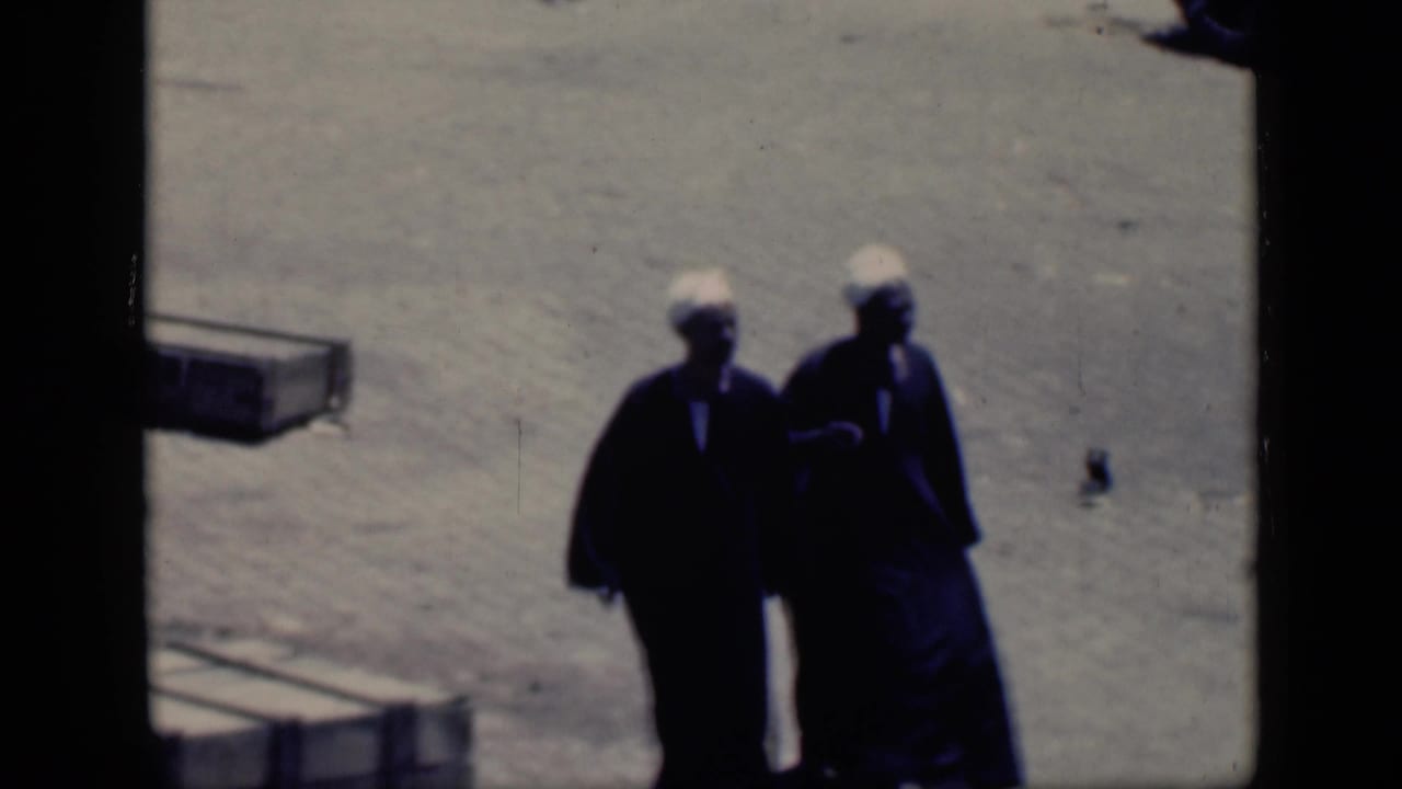 Two adults in dark clothing and white head coverings stand or walk close together in bright sun.