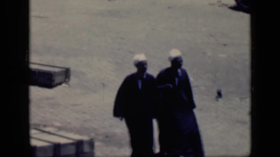 In the Cairo home-movie frame, two adults in dark robes and white head coverings move close together across the open ground.
