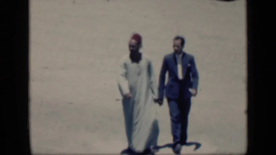 In 1946 Cairo, a man in a light robe and red fez walks hand in hand with a man wearing a dark suit across sunlit ground.